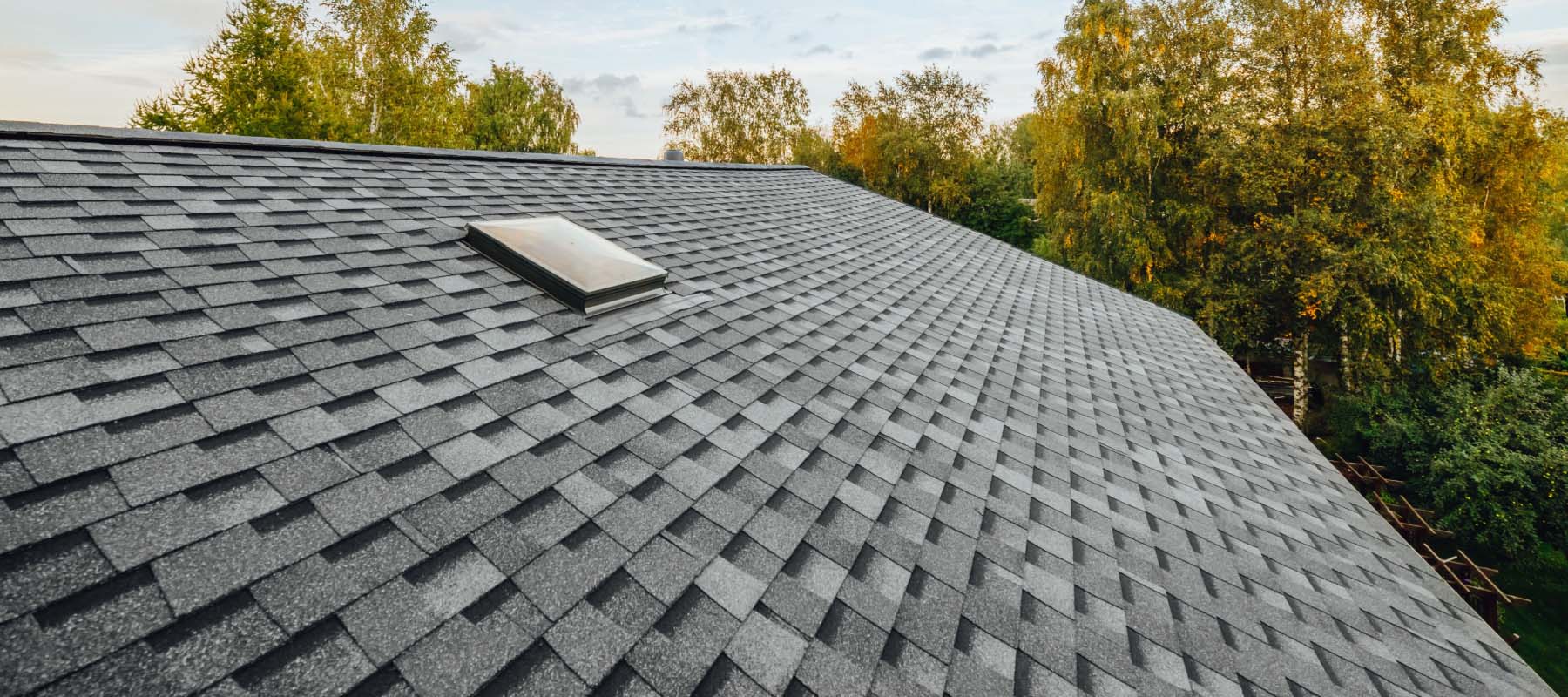 Close-up view of a gray shingled roof with a small skylight, surrounded by green trees and under a partly cloudy sky—an ideal example of quality roofing contractor Suffolk County craftsmanship in NY.