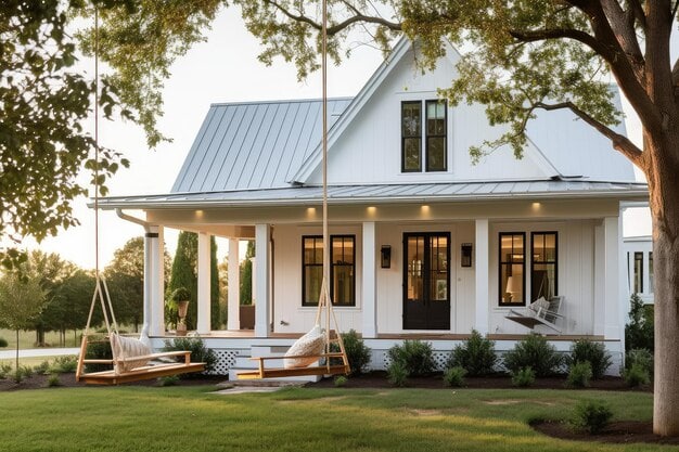 A charming white farmhouse in NY with a metal roof, wide front porch, and two wooden swing benches. Surrounded by green grass and shaded by large trees—perfect inspiration for your next home improvement Suffolk County project.