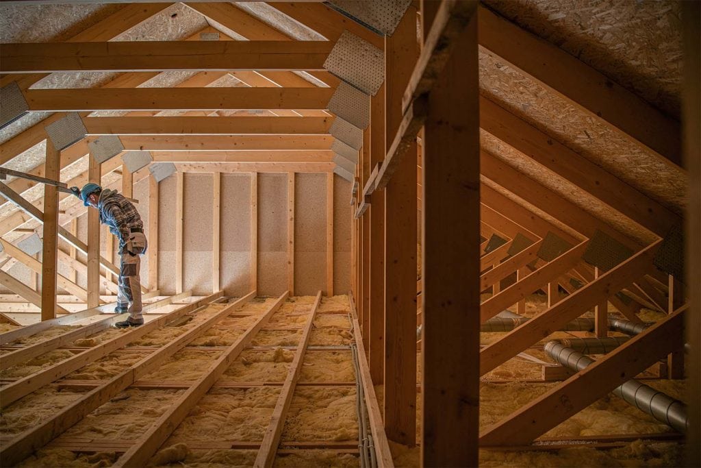 A construction worker wearing safety gear stands in a wooden attic, inspecting the space with exposed beams and insulation on the floor—an example of meticulous home improvement Suffolk County, NY residents can rely on.