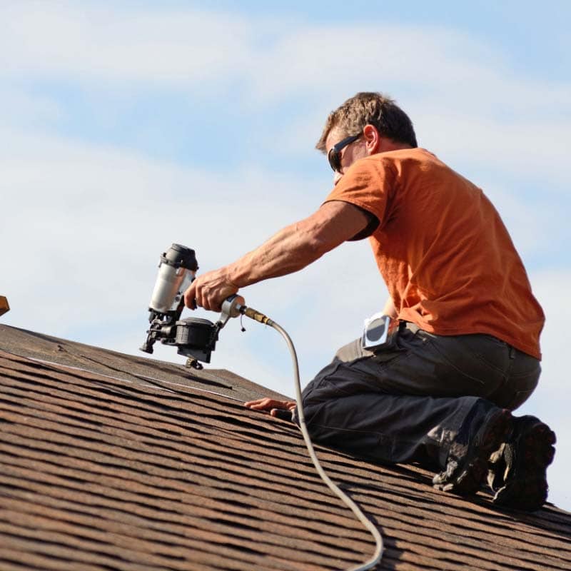 A person in an orange shirt and safety glasses uses a nail gun while working on the shingles of a sloped roof under a blue sky, showcasing skilled home improvement in Suffolk County, NY.