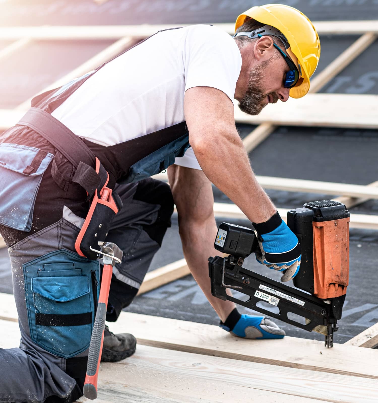 A construction worker in a yellow hard hat and gloves kneels on a wooden roof framework, using a nail gun. He wears safety glasses and has a hammer in his tool belt, representing a skilled roofing contractor Suffolk County, NY.