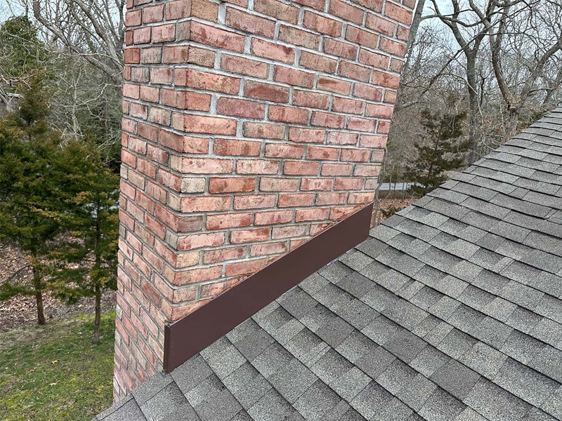 A close-up view of a brick chimney on a shingled roof, showcasing new brown metal flashing installed at the base by a roofing contractor Suffolk County, with trees visible in the background.