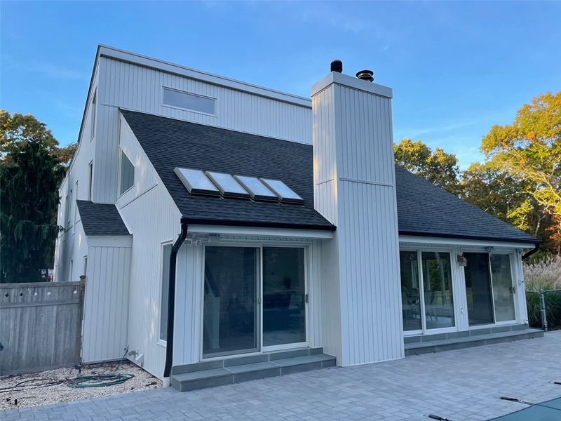 Modern white two-story house with large windows, a slanted black shingle roof by a top roofing contractor Suffolk County, NY, skylights, and a prominent exterior chimney, surrounded by trees and stone pavement.