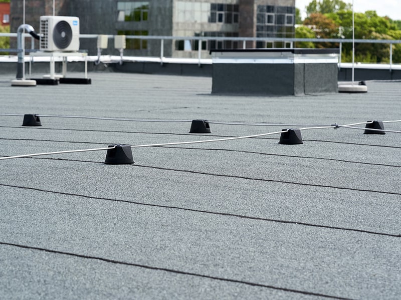 Flat building rooftop with black safety cones connected by a wire, an air conditioning unit, and a raised rectangular structure; ideal for inspection by a roofing contractor Suffolk County. City buildings and trees are visible in the background.