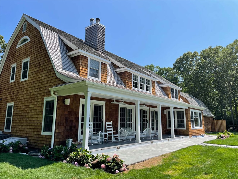 A large, two-story wooden house with a shingled roof by a trusted roofing contractor Suffolk County, featuring dormer windows, a spacious covered patio with white rocking chairs and garden furniture, surrounded by green grass and trees.