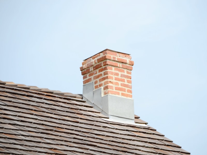 A red brick chimney with metal flashing is built on a brown shingled roof against a clear, pale blue sky, showcasing the quality work of a roofing contractor Suffolk County.