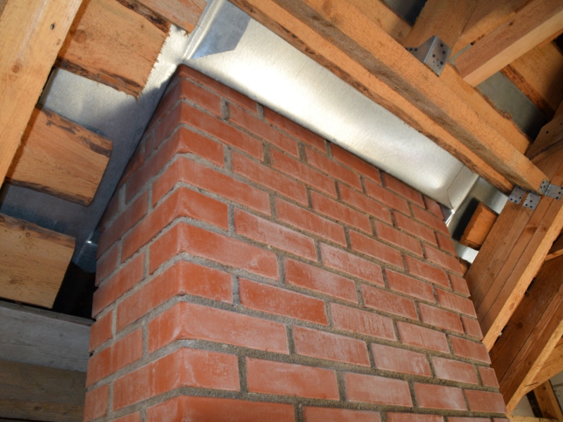 A close-up view of a red brick chimney passing through a wooden roof structure, with metal flashing installed for waterproofing by a skilled roofing contractor Suffolk County, NY.