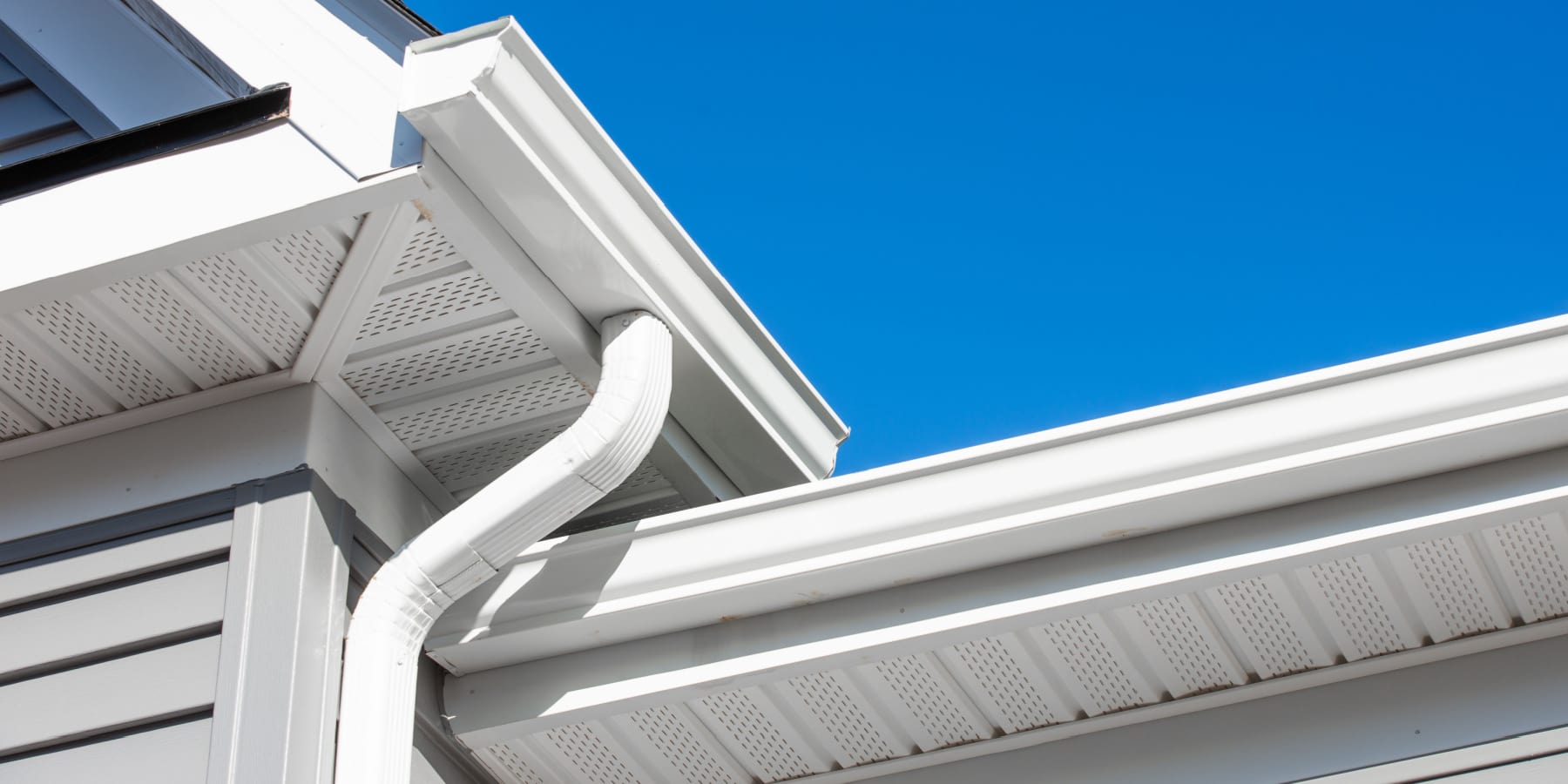 White rain gutter and downspout system attached to the corner of a house with light gray siding, set against a clear blue sky—ideal for your next home improvement Suffolk County, NY project.