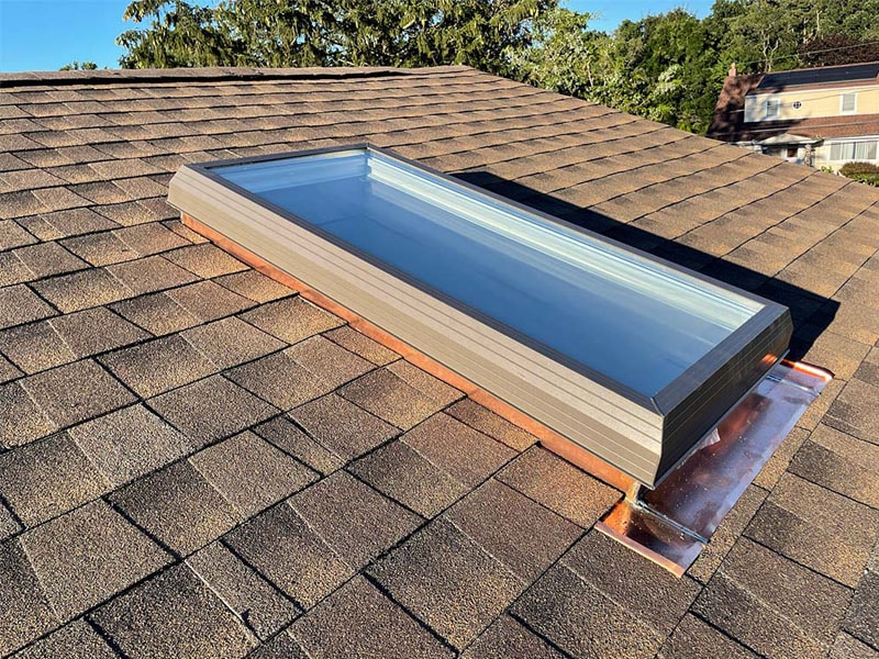 A rectangular skylight with a metal frame installed on a sloped, brown shingle roof by a roofing contractor Suffolk County; sunlight reflects off the glass, with nearby houses and trees visible in the background.