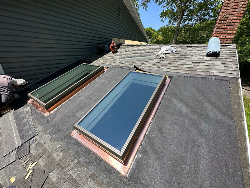 Two workers from a roofing contractor in Suffolk County, NY, install new skylights on a shingled roof. Surrounded by copper flashing and tools, the home improvement project is set against trees and a brick chimney in the background.