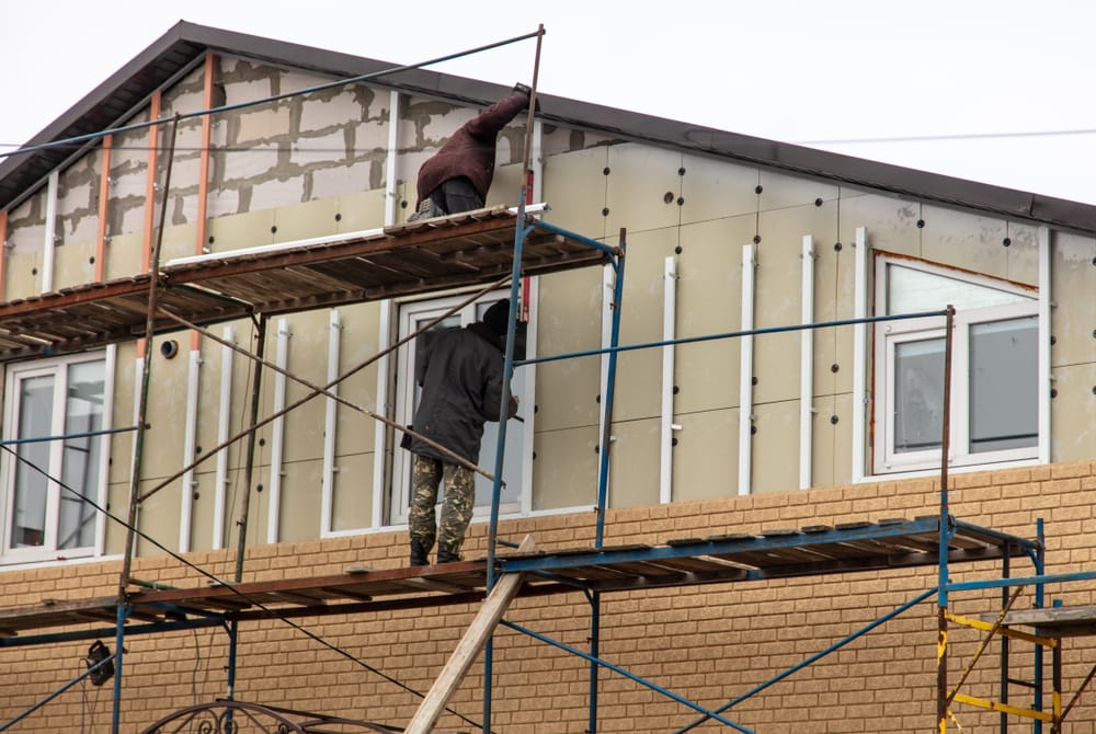 Two workers on scaffolding install exterior insulation panels on the upper floor of a house under renovation—a typical scene for a home improvement Suffolk County, NY project, with exposed insulation and some completed brickwork on the lower wall.