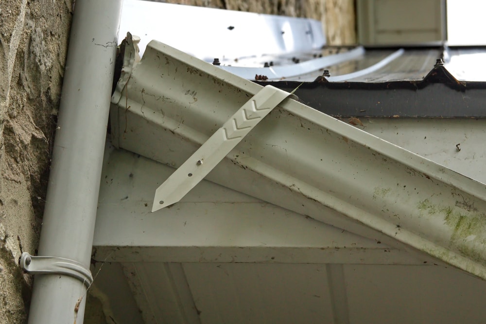 A broken and bent white metal bracket hangs loosely from a dirty, weathered rain gutter on a stone building—an issue that could benefit from the expertise of a roofing contractor Suffolk County, NY.