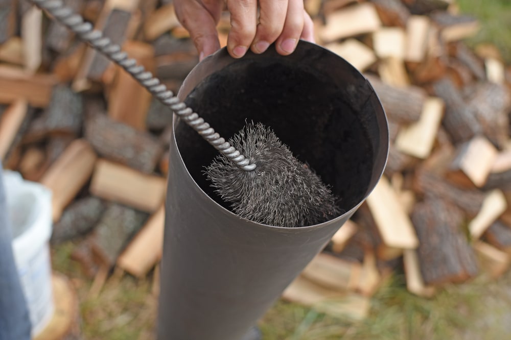A hand holds a twisted wire chimney cleaning brush inside a metal pipe, with chopped firewood scattered on the ground—perfect for any home improvement Suffolk County, NY project needing attention to chimney maintenance.