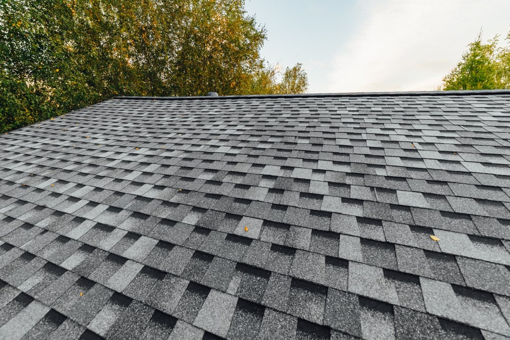 Gray asphalt shingle roof with a pattern of overlapping tiles, surrounded by green trees under a partly cloudy sky—perfect inspiration for your next home improvement Suffolk County project.