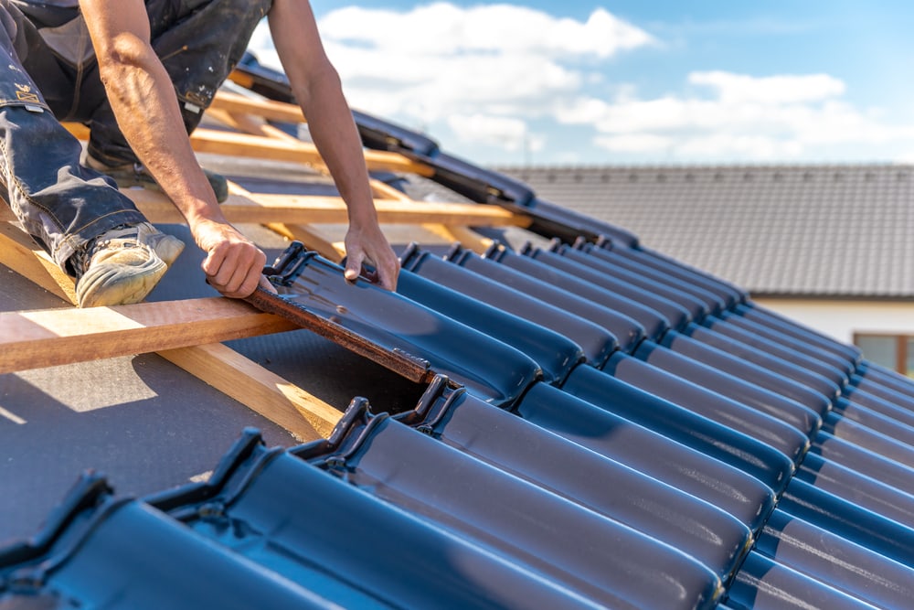 A construction worker, part of a roofing contractor Suffolk County team, installs dark blue roof tiles on a wooden house frame under a bright sky, with several tiles already in place.