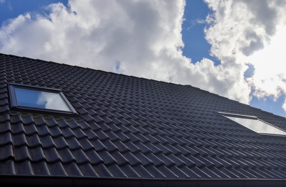 A sloped roof with dark, glossy tiles and two skylight windows, set against a partly cloudy blue sky—an inspiring example by a roofing contractor Suffolk County, NY.