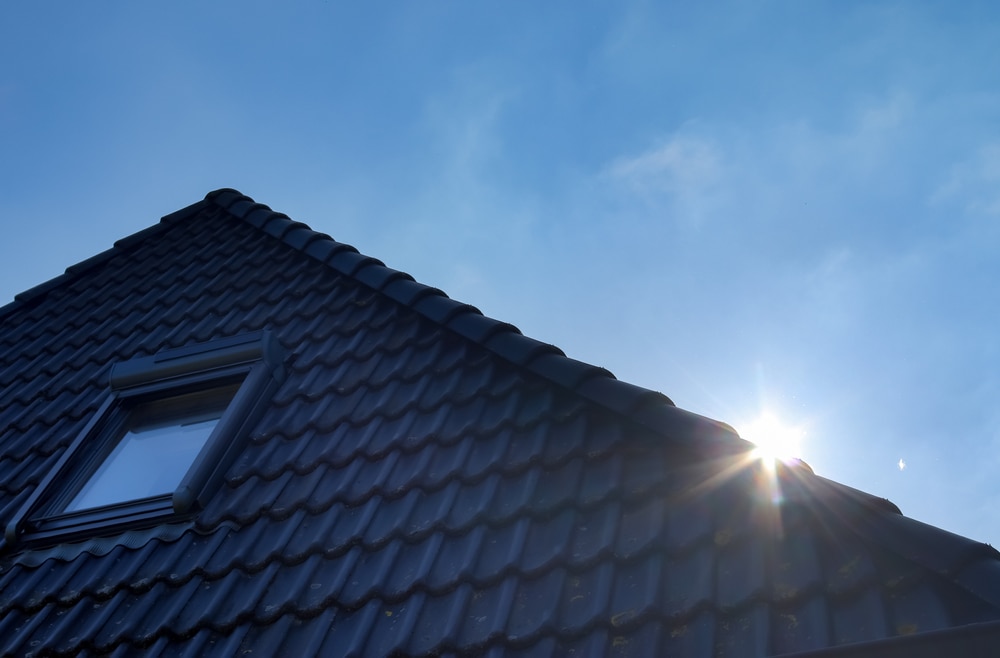 A close-up view of a dark tiled house roof with a single window, sunlight shining at the corner against a clear blue sky—perfect inspiration for your next home improvement Suffolk County project.