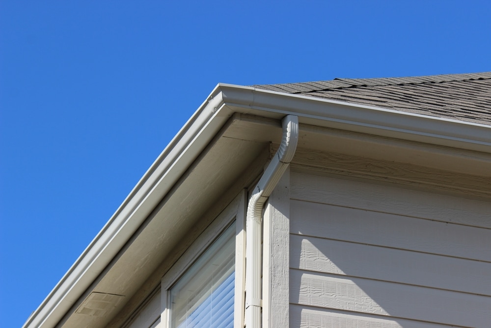 Close-up view of the corner of a house with beige siding, white rain gutter, and downspout under a shingled roof against a clear blue sky—perfect inspiration for your next home improvement Suffolk County, NY project.