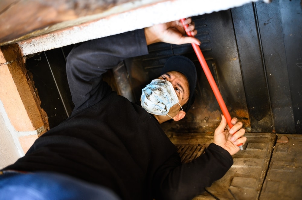 A person wearing a cap and a protective face mask lies on the ground, working under a fireplace with a red tool, appearing to perform maintenance—possibly part of a home improvement Suffolk County project.