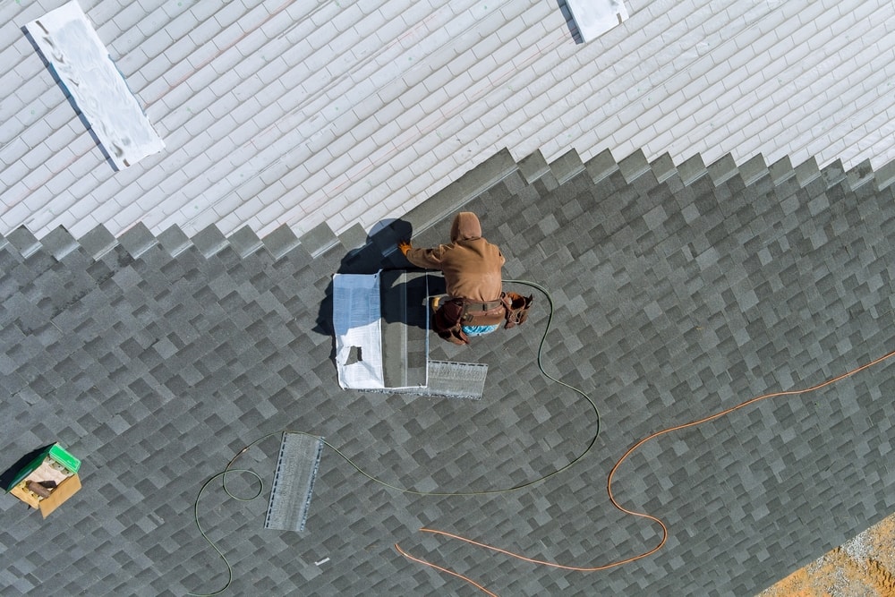 A person in a brown hoodie installs or repairs a vent on a gray shingled roof in NY, with tools and materials nearby. The image is taken from above, showing a clear view of the roof and equipment, ideal for home improvement Suffolk County projects.