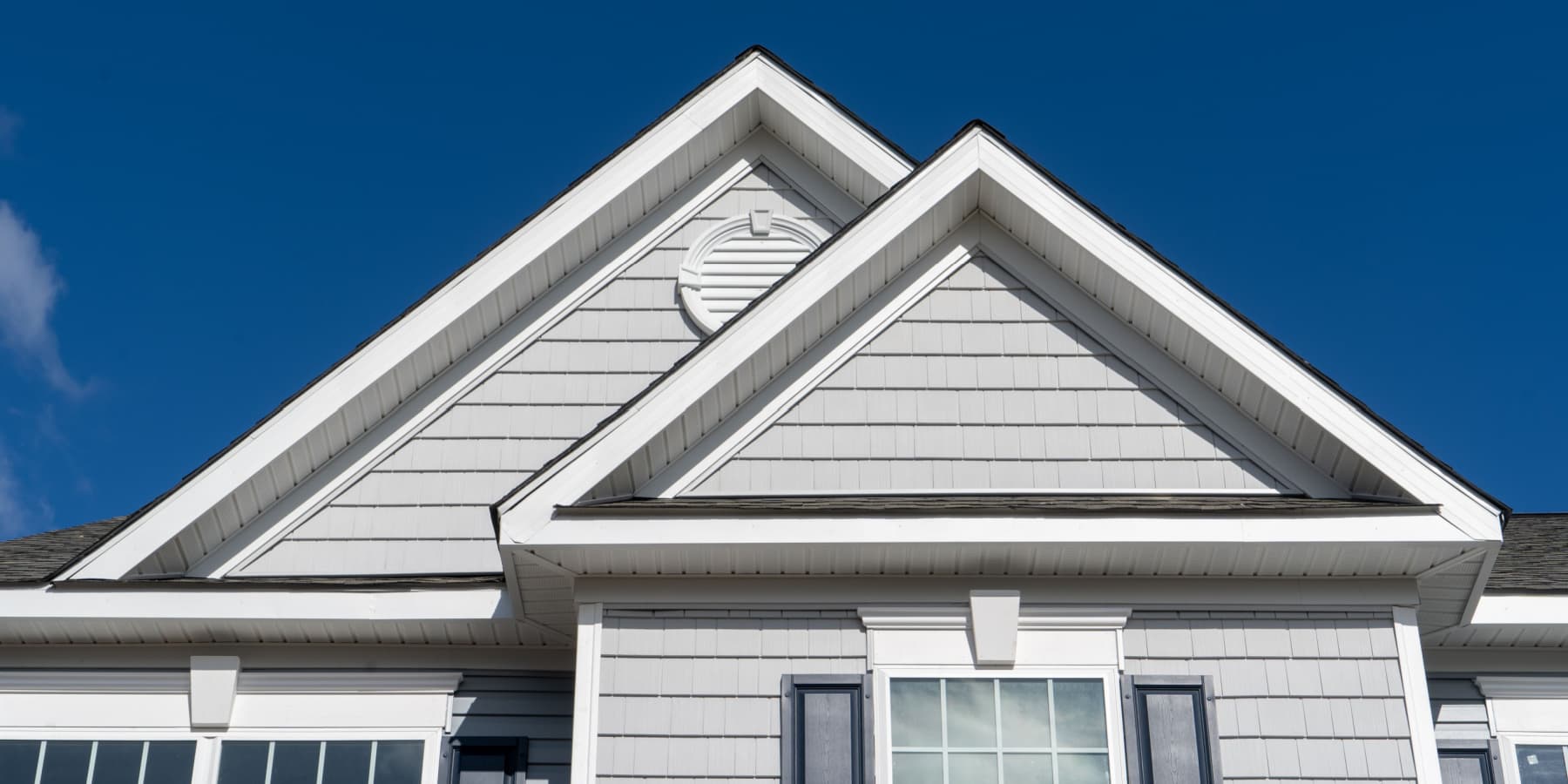 Close-up of the upper front of a gray house with white trim, featuring two prominent gables against a clear blue sky. Ideal inspiration for home improvement Suffolk County, NY, with navy shutters and a circular vent completing the look.