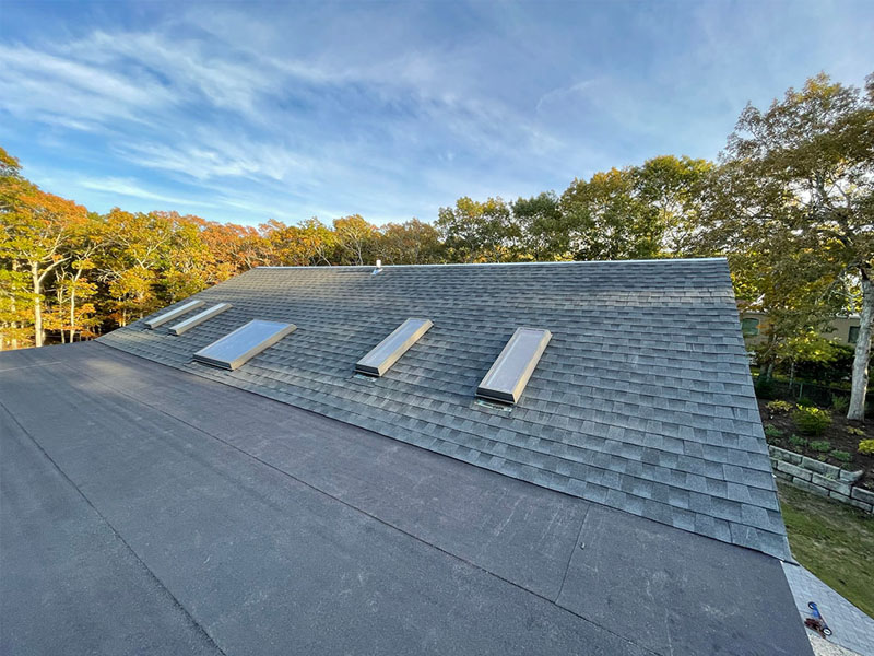 A sloped roof with four rectangular skylights, covered in gray shingles, surrounded by trees with green and autumn-colored leaves under a blue NY sky—perfect inspiration for your next home improvement Suffolk County project.
