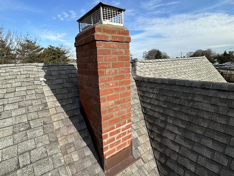 A red brick chimney with a metal cap stands on a gray shingled roof under a partly cloudy sky in NY, with trees visible in the background.