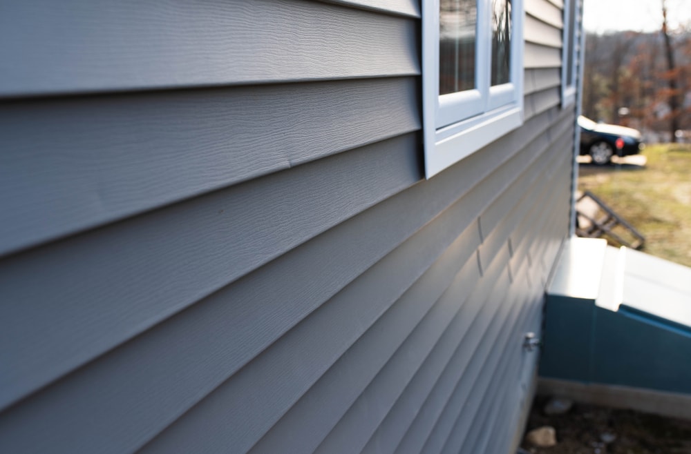 Close-up view of gray vinyl siding on a home's exterior wall, with a window and part of a stairway visible; a blurred car and yard appear in the background—perfect inspiration for your next home improvement in Suffolk County, NY.