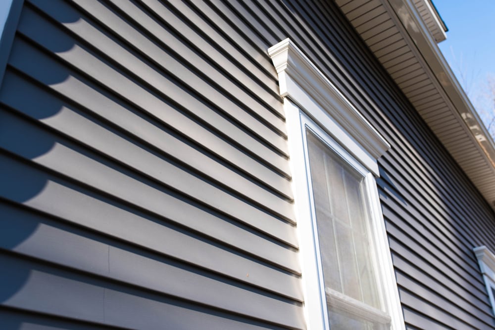 Close-up view of gray vinyl siding and a white-trimmed window in sunlight, highlighting details ideal for any home improvement Suffolk County, NY project.