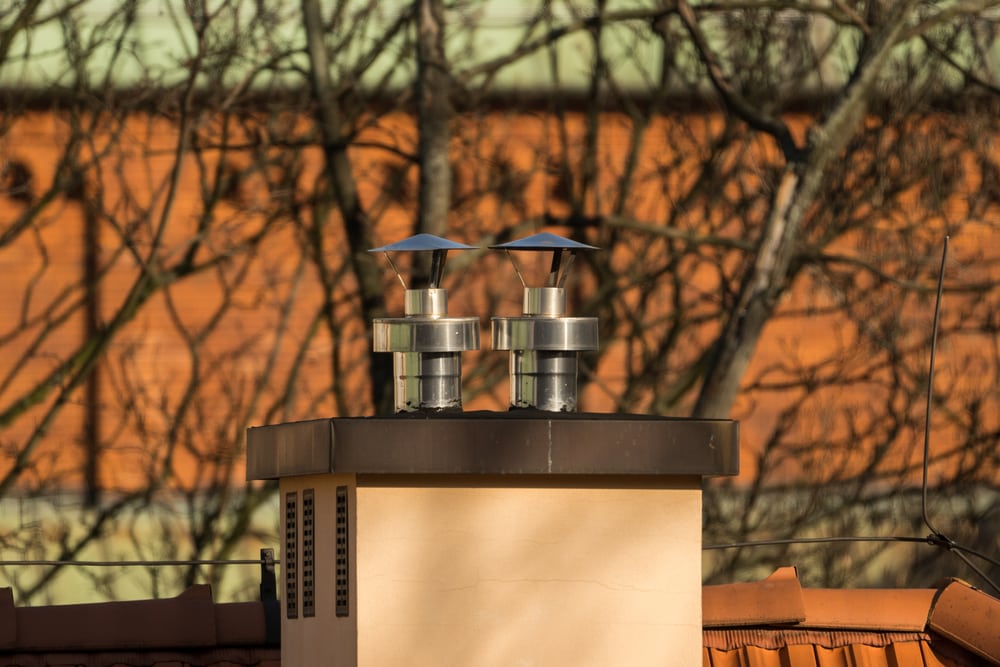 Two metal chimney caps sit atop a beige chimney structure—an ideal detail for any home improvement Suffolk County project—with bare tree branches and a blurred brick building in the background.