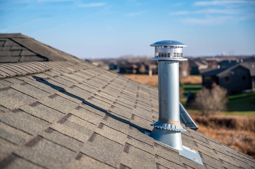 A metal chimney vent pipe extends from the sloped shingle roof of a house, expertly installed by a roofing contractor Suffolk County, with houses and grassy fields visible in the blurred background under a blue sky.