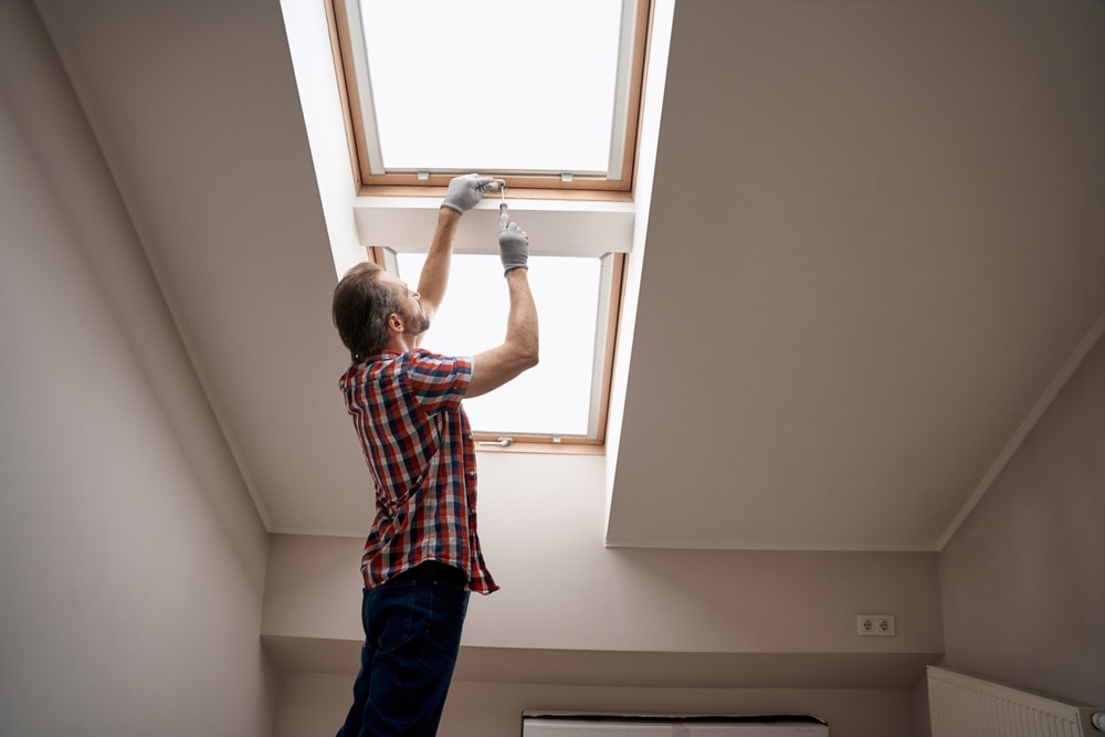 A man wearing gloves and a plaid shirt stands on tiptoe inside a room, adjusting a skylight window on the slanted ceiling. Natural light comes through the window—an example of home improvement Suffolk County, NY homeowners may need.