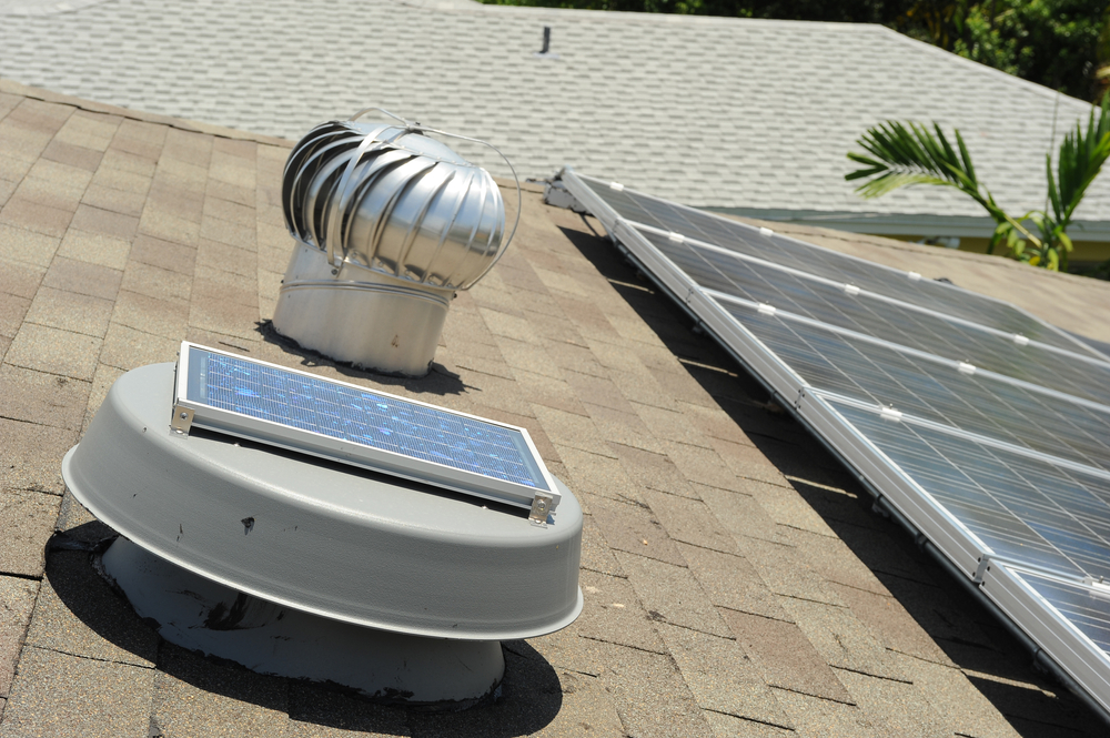 A metal turbine vent and a small solar panel are installed on a shingled roof, showcasing quality work by a roofing contractor Suffolk County, with a larger array of solar panels and a palm tree visible in the background.