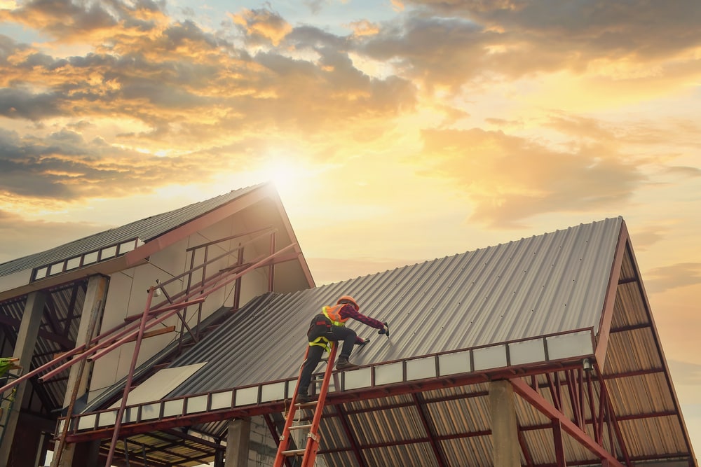 A construction worker on a ladder installs metal roofing panels on a building at sunset, with sunlight shining through a partly cloudy sky—a skilled roofing contractor in Suffolk County, NY, enhancing home improvement projects.