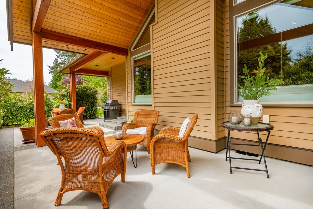 Outdoor patio with wicker chairs and a wooden table under a covered area attached to a modern beige NY home. A small side table holds a potted plant and pitcher, while a barbecue grill sits nearby—perfect for your next home improvement Suffolk County project.