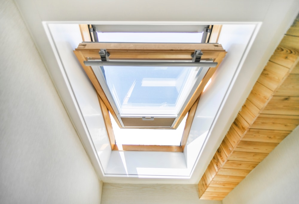 A wooden-framed skylight window is open in a white ceiling, letting sunlight stream in—an ideal home improvement for Suffolk County, NY. Wooden steps or a ladder are visible on the right side, leading up to the window.