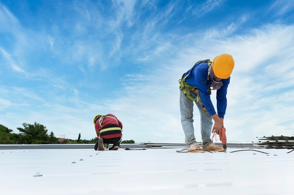 Two construction workers in safety gear and helmets install or repair a metal roof under a bright blue sky. Perfect for those seeking home improvement in Suffolk County—trust a skilled roofing contractor Suffolk County for quality results.