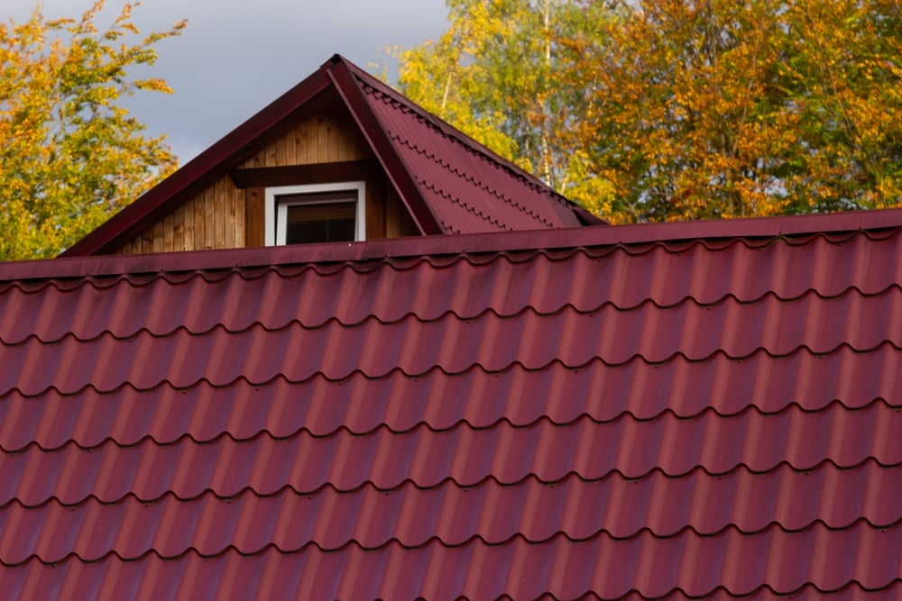 A house with a red metal tile roof and a small dormer window, expertly crafted by a roofing contractor Suffolk County. Trees with green and yellow autumn leaves provide a stunning NY backdrop.