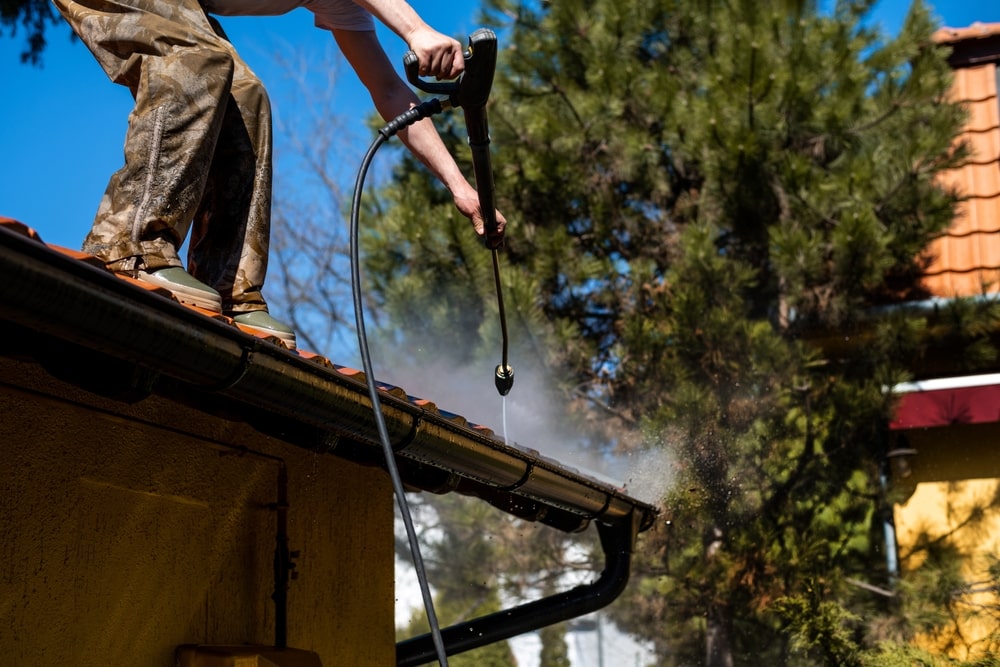 A person stands on a roof, using a pressure washer to clean the gutters—a great example of home improvement Suffolk County, NY style. Water sprays from the nozzle, and trees and a house can be seen in the sunny background.
