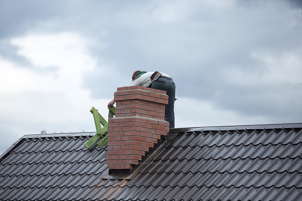 A roofing contractor Suffolk County works on a steep, metal-tiled roof next to a brick chimney under a cloudy NY sky, using a green ladder for support.