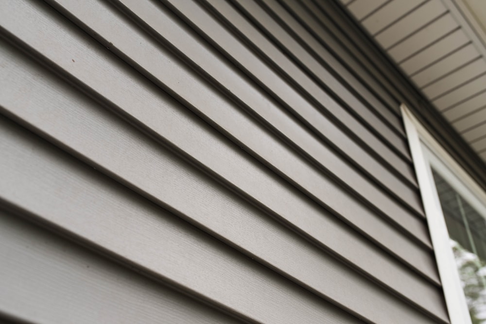 Close-up view of the exterior wall of a house with horizontal beige vinyl siding, a white-trimmed window on the right, and part of the roof overhang installed by a skilled roofing contractor Suffolk County at the top.