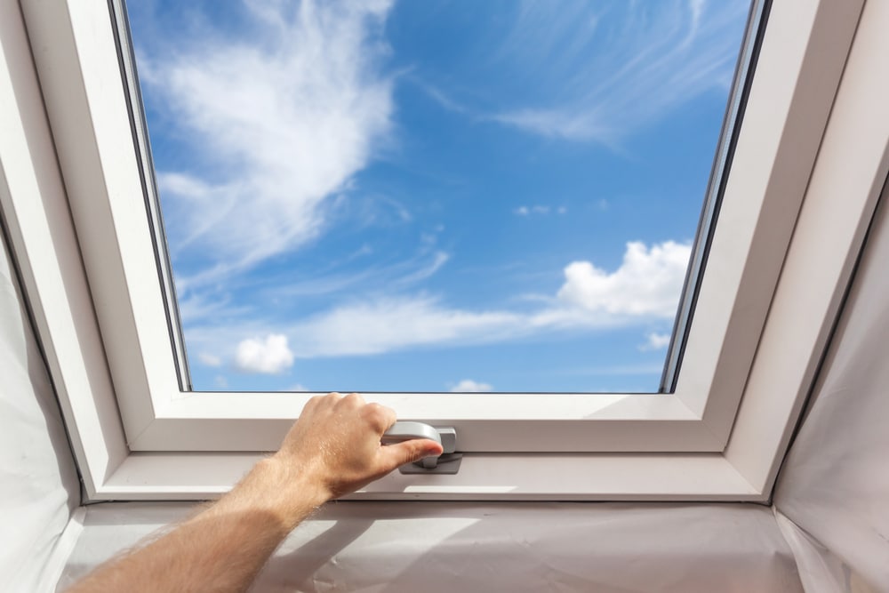 A person’s hand opens a white-framed skylight window, revealing a bright blue sky with scattered clouds—sunlight pours in, showcasing the work of a skilled roofing contractor Suffolk County and inspiring home improvement Suffolk County dreams.