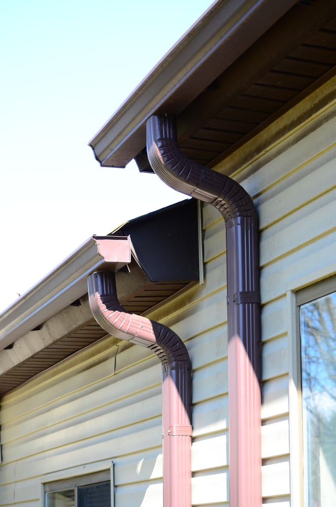 Two brown downspouts attached to the side of a yellow house, running from the roof gutter downward; sunlight casts shadows on the siding—a great example of detail for any home improvement Suffolk County project.