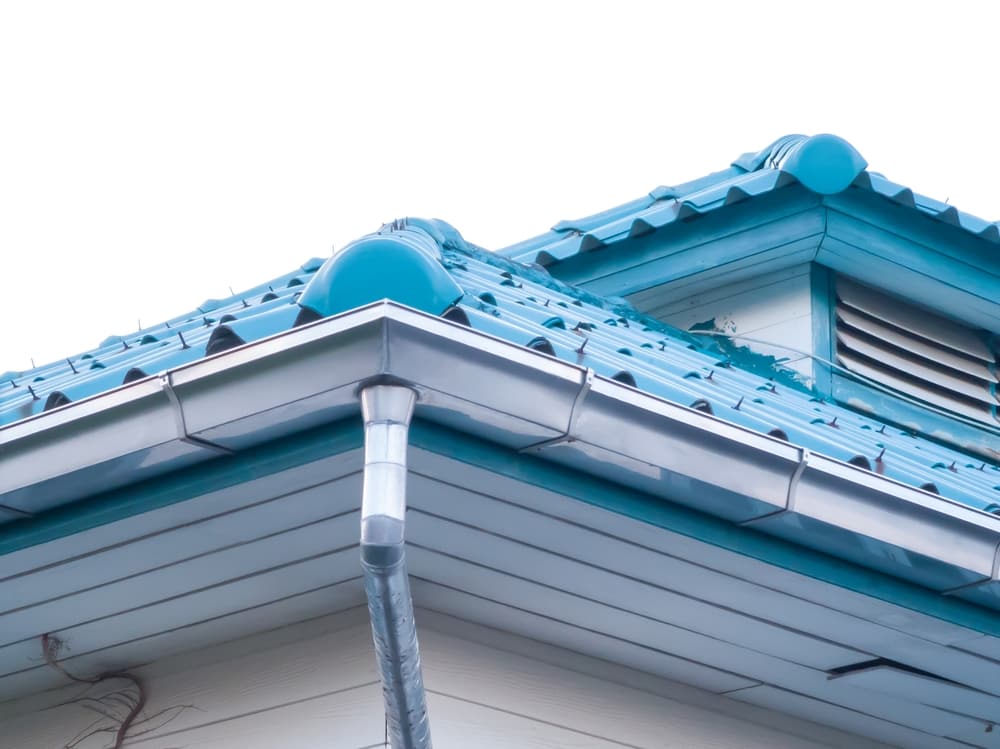 Close-up view of a house’s blue tiled roof with a silver gutter and downspout, white siding, and a louvered vent on the upper wall—an example of expert work by a roofing contractor Suffolk County, set against a bright, overexposed NY sky.