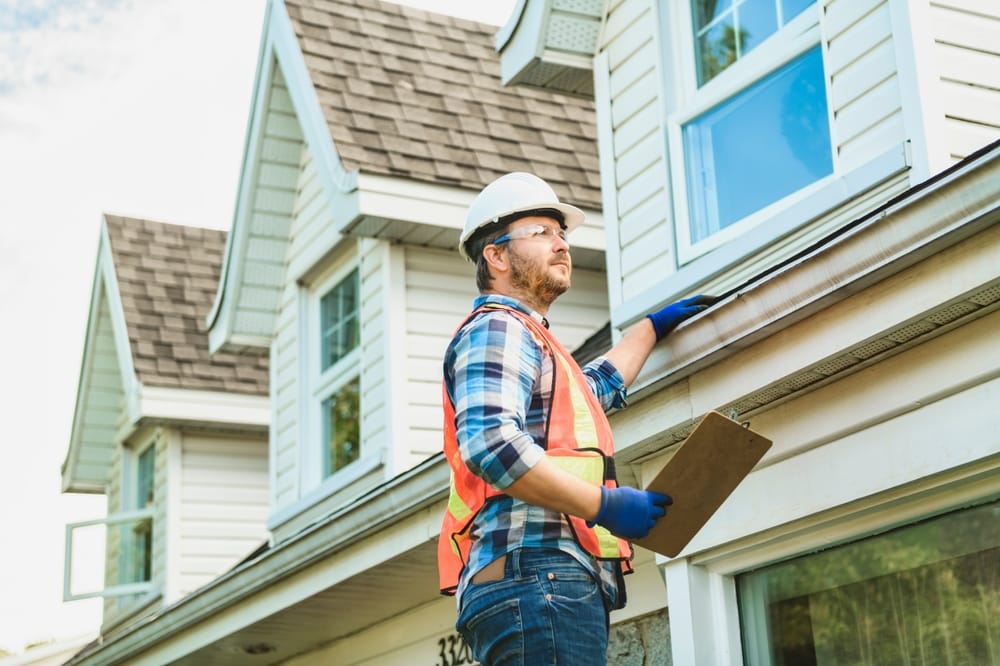 A construction worker wearing a hard hat, safety vest, and gloves inspects the roof and gutter of a house while holding a clipboard. The home improvement Suffolk County, NY project features white siding and multiple dormer windows.