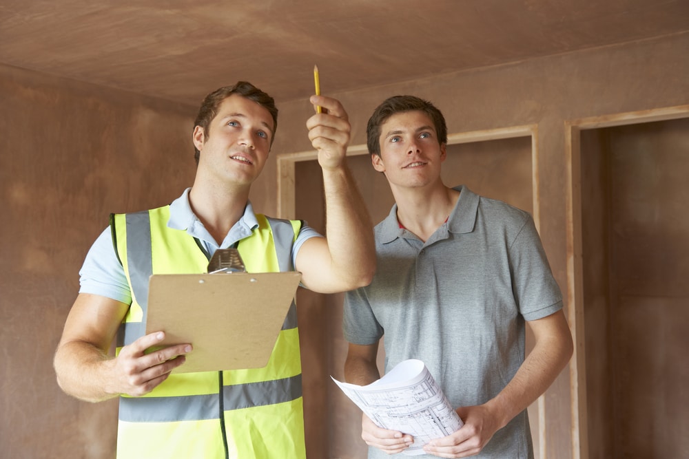 Two men stand in an unfinished room; a roofing contractor Suffolk County in a reflective vest gestures toward the ceiling with a pencil, while the other holds blueprints and looks in the same direction.