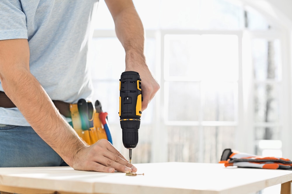 A person using a yellow and black cordless power drill to drive a screw into a wooden surface; tool belt with tools visible, sunlight streaming through large windows—perfect for any NY home improvement Suffolk County project.