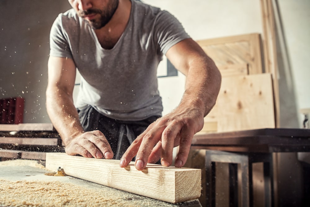 A man in a gray t-shirt sands a wooden plank on a workbench, surrounded by wood dust and tools—capturing the spirit of hands-on home improvement Suffolk County, NY is known for.
