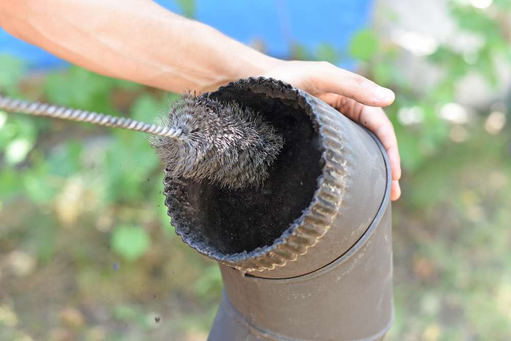 A person holds a metal pipe while cleaning its interior with a cylindrical wire brush. The background is blurred with greenery visible, suggesting a NY home improvement Suffolk County project in progress.