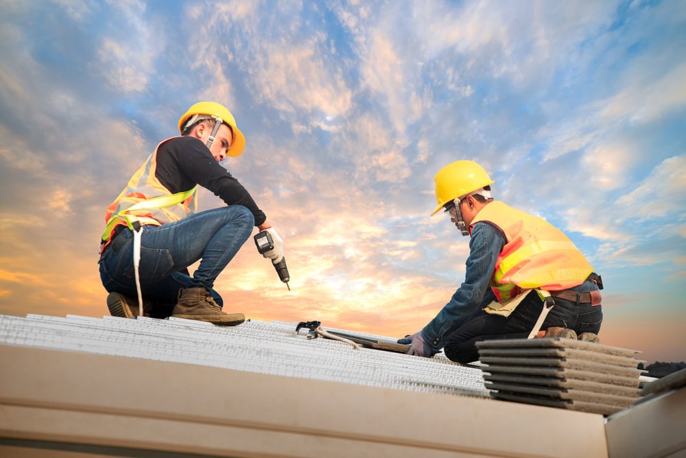 Two construction workers in safety vests and helmets install roofing material on a building at sunset in NY, representing a skilled roofing contractor Suffolk County, using power tools and surrounded by roofing tiles.